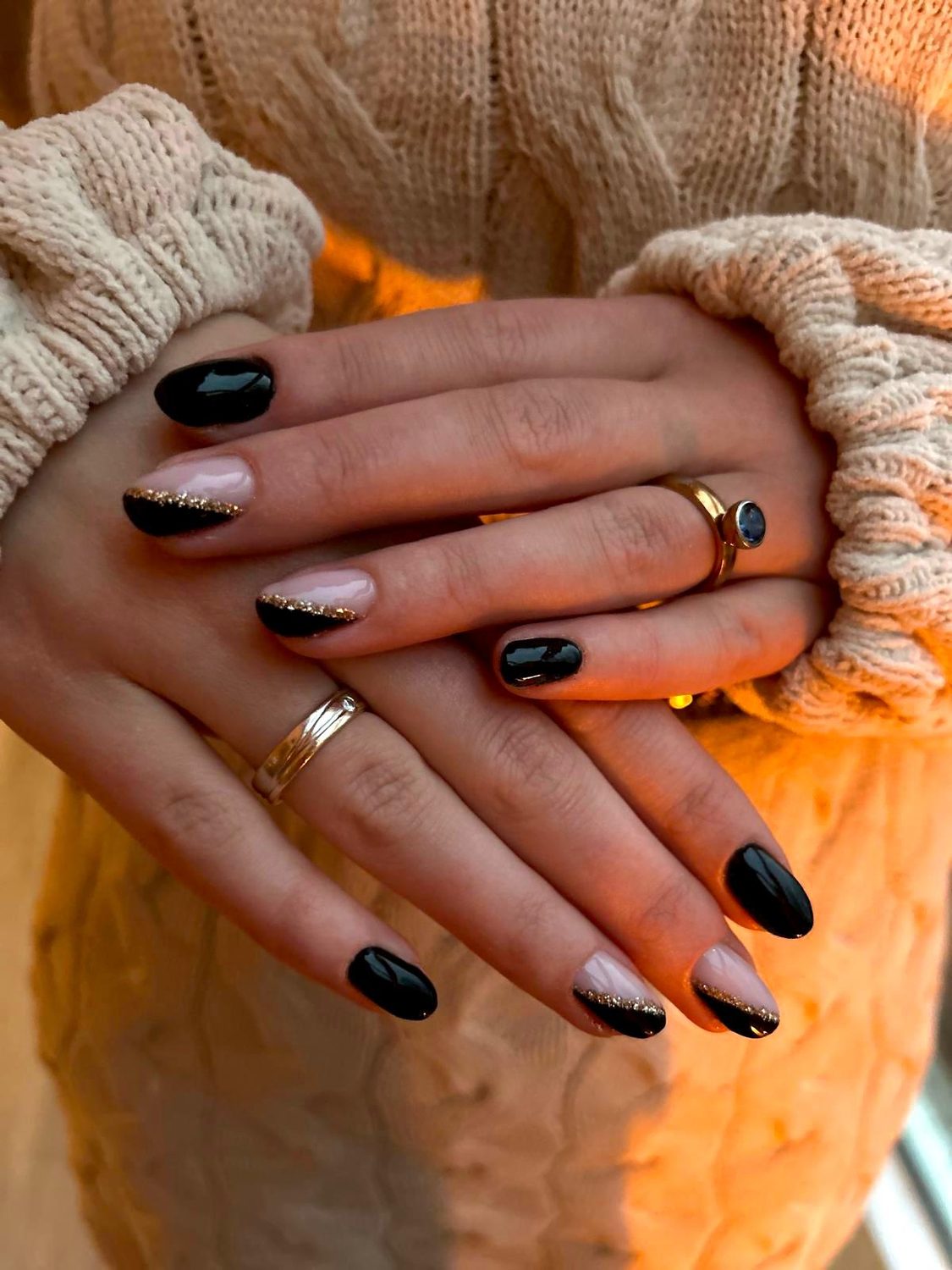 Zwarte en nude nagels met een fijne glitterlijn, handen gevouwen voor een oranje gebreide trui